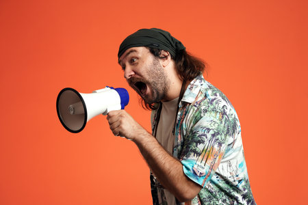 A man holds a megaphone and speaks loudly into it, shouting. Male traveler in the studio on an orange background close up. Concept of travel.の写真素材