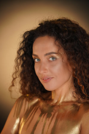 Portrait of female model in studio. Attractive young woman with curly hair and golden makeup with covered chest in golden paint looks at camera.の写真素材