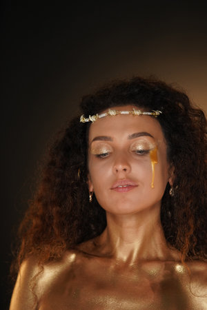 Portrait of female model in studio. Attractive girl wearing tiara and makeup covered in golden paint posing at camera with golden tear on cheek.の写真素材