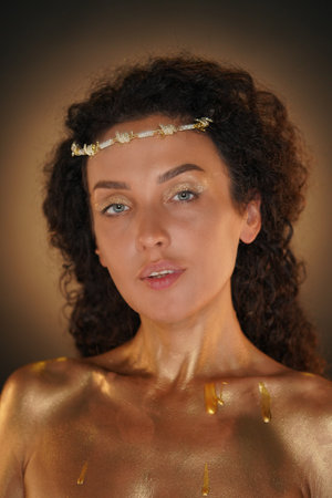 Portrait of female model in studio. Close up shot of attractive girl wearing tiara and elegant makeup body covered in golden paint looking at camera.の写真素材