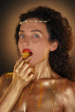 Portrait of female model in studio. Attractive girl wearing tiara and red lipstick, body covered in golden paint holding golden candy in mouth.の写真素材