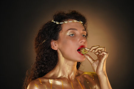 Portrait of female model in studio. Appealing girl wearing tiara and red lipstick, body covered in golden paint holding golden strawberry in lips.の写真素材
