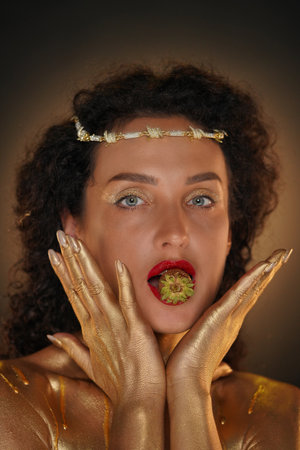 Portrait of female model in studio. Appealing girl in tiara covered in golden paint holds strawberry in lips, hand touching face. looks at camera.の写真素材
