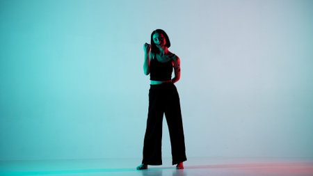 In the frame on a blue, light background. Dances young, beautiful girl. Demonstrates dance moves in the style of hip hop. Shes staring at the camera. Shes feminine in a black top and pantsの写真素材