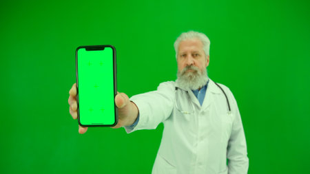 Portrait of man medic in studio on chroma key green screen. Senior doctor in white uniform holding smartphone with workspace mockup, close up shot.の写真素材