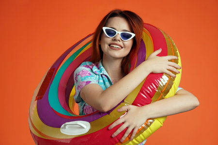 Close up of woman in glasses hugging colorful inflatable pool tube and smiling at the camera, isolated on orange background. Portrait of female traveler.の写真素材