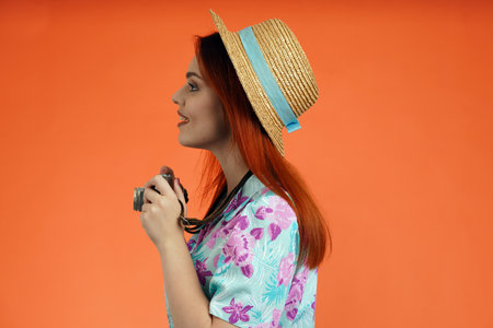 Close up profile shot of woman with old film camera, looking at the side and ready to take pictures, isolated on orange background. Portrait of female traveler.の写真素材
