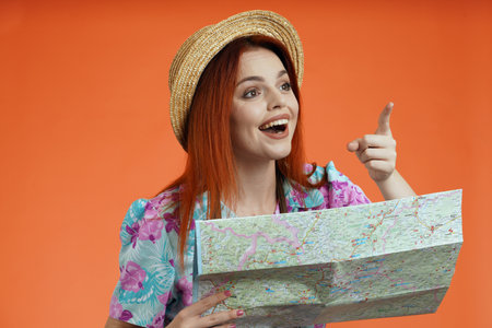 Close up of woman in straw hat smiling, holding paper map pointing finger at the side, found direction, isolated on orange background. Portrait of female traveler.の写真素材