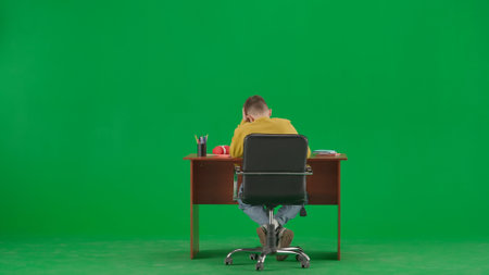 Portrait of kid boy on chroma key green screen. Schoolboy in jeans sitting at the desk doing homework, writing in notebook, reading. Back full shot.の写真素材