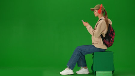 Portrait of person tourist isolated on chroma key green screen background. Young woman sitting holding smartphone listening music in headphones.の写真素材