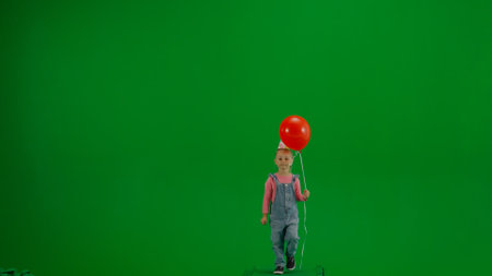 A little girl in denim overalls with a festive cap on her head and carrying a red helium balloon walks in front of a green screen, front view. Concept of a holiday, birthday party.の写真素材