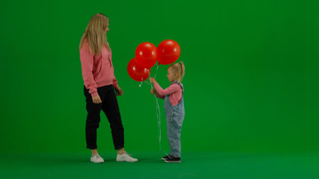 Little girl standing with closed eyes, mom brings her balloons, girl is happy and surprised on chroma key green screen background.の写真素材