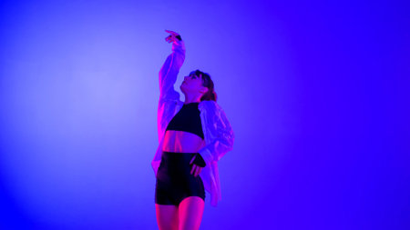 Young woman wearing a top, shorts and a shirt performing contemporary dance in studio. Neon blue,...
