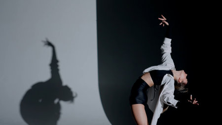 Young woman wearing a top, shorts and a shirt performing contemporary dance under a spotlight in the studio. Contrasting black falling shadow on the background. Medium full.の写真素材