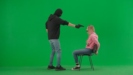 Portrait of thief and victim on chroma key green screen background. Girl sitting on chair tied hands and taped mouth man robber with gun threatens her.の写真素材