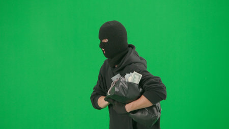 Portrait of thief on chroma key green screen background. Man robber wearing hoodie, jeans and balaclava, running from police with stolen bag full of money. Side view.の写真素材