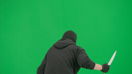 Portrait of thief on chroma key green screen background. Man robber in balaclava and hoodie walking holding knife in hand looks around. Back view.の写真素材