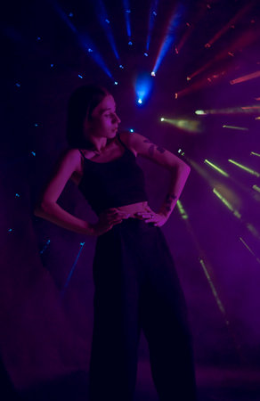 A young woman in black casual clothes poses in a studio with smoke, purple light and beams of multicolored light. The dancer demonstrates elements of experimental hip hop style dance choreography.の写真素材