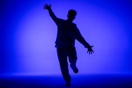 Silhouette of guy in casual clothes dancing elements of hip hop in studio with blue light. Dancer demonstrates body plasticity. Full height. Modern street choreography.の写真素材