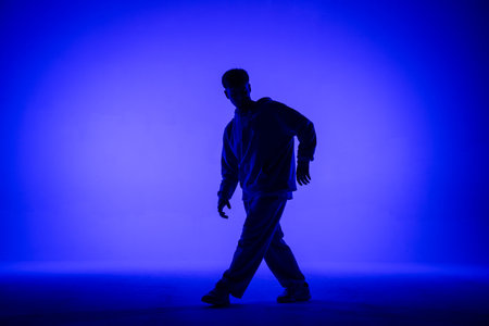 Silhouette of guy in casual clothes dancing elements of hip hop in studio with blue light. Dancer demonstrates body plasticity. Full height. Modern street choreography.の写真素材