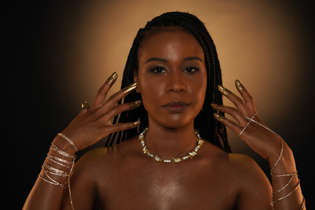 Portrait of a young African American woman with gold jewelry. The models arms are entwined with gold chains, and there is a necklace around her neck. Liquid gold is applied to the fingers and nails.の写真素材