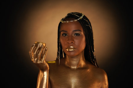 Portrait of an African-American woman with gold jewelry in her hair. The models arms, neck and shoulders are covered with liquid gold. The girl is holding a golden lemon in her hands. Close upの写真素材