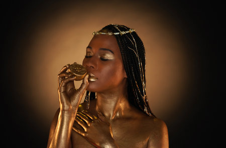 Portrait of an African-American woman with gold jewelry in her hair. The models arms, neck and shoulders are covered with liquid gold. The girl is holding a golden lemon in her hands. Close upの写真素材