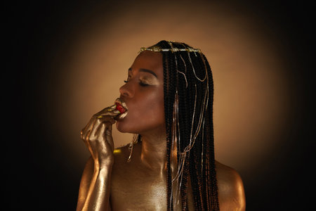 Portrait of an African-American woman with gold jewelry in her hair. The models arms, neck, lips and shoulders are covered with liquid gold. A girl is biting a strawberry. Close up.の写真素材