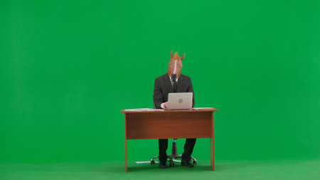 Man in business suit with horse head mask on studio green background. Businessman sitting at desk and typing on laptop keyboard. Heavy office work concept.の写真素材