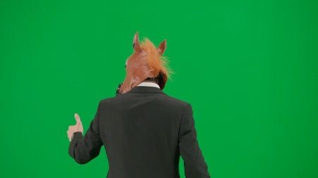 Man in business suit with horse head mask on green studio background. Businessman walking, pointing fingers in the direction and showing thumbs up gesture. Concept of hard office work. Back view.の写真素材