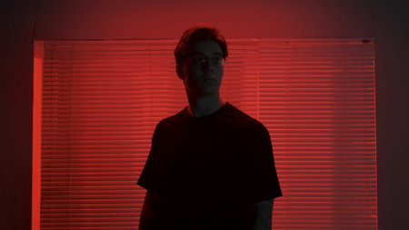 Portrait of male in dark room. Handsome man near window, red neon light shines behind jalousie, guy looks away naturally with calm expression.の写真素材