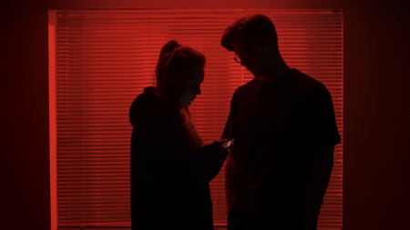 Portrait of male in dark room. Handsome man near window, red neon light shines behind jalousie, girl next to man checks smartphone.の写真素材