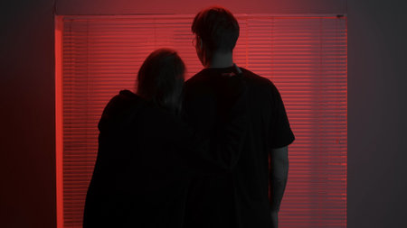 Portrait of male in dark room. Handsome man and girl standing hugging facing the window, red neon light shines behind jalousie.の写真素材