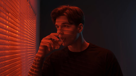 Portrait of male in dark room. Handsome man near window, red neon light shines behind jalousie, guy looks outside drinks whiskey from a glass.の写真素材