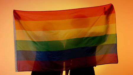 Silhouettes of people with large flag on yellow background. Silhouettes of a man and a woman with LGBT pride flag holding in hands.の写真素材