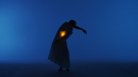 Female dancer performing in the studio. Young woman dancing in dark studio with warm light glowing inside the chest under her shirt.の写真素材
