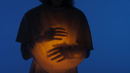 Female dancer performing in the studio. Young woman in dark studio touching warm light glowing inside the chest under her shirt, close up.の写真素材