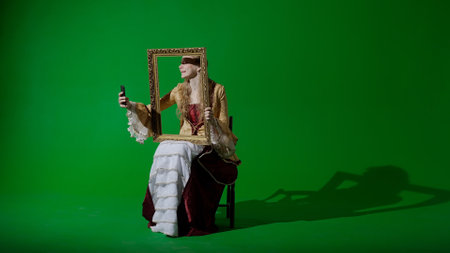 Woman in ancient outfit on chroma key green screen background. Female in renaissance style dress holding painting frame taking selfie on smartphone.の写真素材