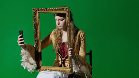 Woman in ancient outfit on chroma key green screen background. Female in renaissance style dress holding painting frame taking selfie on smartphone.の写真素材