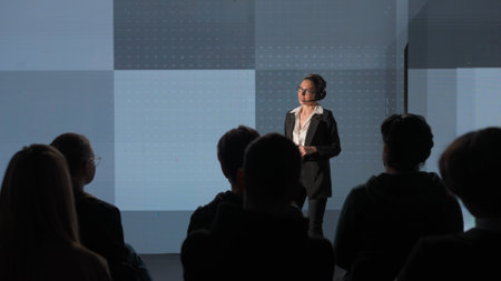 Woman presenting project on stage during meeting. Shot from audience, people sitting listening businesswoman speaker talks shows data analytics infographic on a huge LED screen.の写真素材