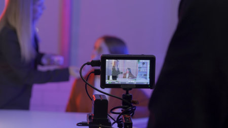 Camera on a blurred background of a female makeup artist applying blush to the face of a female presenter. Film crew in a dark studio in pink neon light.の写真素材