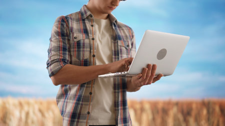 Farmer against wheat field and blue sky. Man agronomist standing in the farm field, holding laptop...