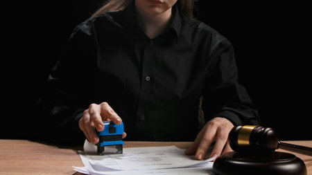 Female sitting at the desk with wooden hammer, dark room bright spotlight. Woman judge silhouette in the court room, looking through documents, reading papers placing stamp.の写真素材