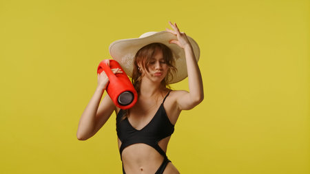 Young woman in swimsuit and stylish hat with music speaker dances lively at camera. Travel, summertime vacation, beach leisure concept.の写真素材