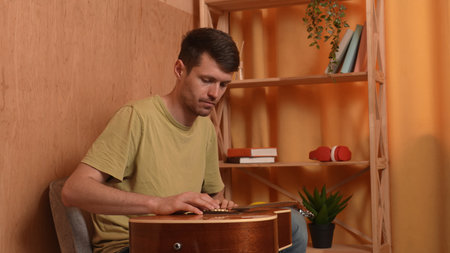 Adult man guitarist sitting in the room playing acoustic guitar tapping on wooden body part,...
