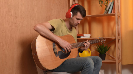 Adult man guitarist sitting in the room playing acoustic guitar, listening music in headphones, composing fingering chords.の写真素材