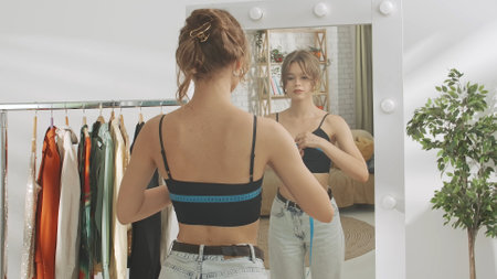Young millennial woman in her bedroom looks in the mirror, measures her chest size, upset disappointed face. Self care healthy beauty concept.の写真素材