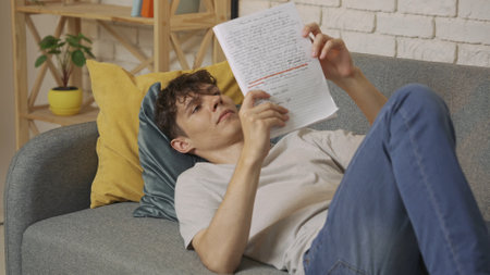 Focused adult man college student laying on sofa in living room at daylight and doing homework, holding notebook reading essay. Education concept.の写真素材