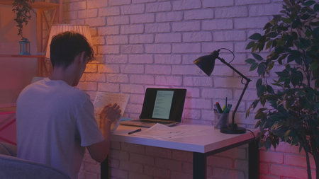 Focused adult man college student sitting at desk in apartment room late at night and doing homework...