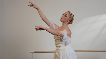 A beautiful ballerina in a white dress dances in a white sunny room next to a ballet barre.